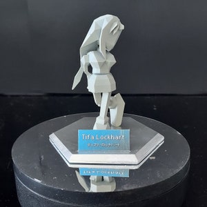 Op de afbeelding: Een wit, low-poly beeldje van een vrouwelijk personage met lang haar, dat een kort rokje en laarzen draagt. Het beeldje staat op een ronde, zilveren voet met een blauw naamplaatje waarop "Tifa Lockhart" en "ティファ・ロックハート" staat.