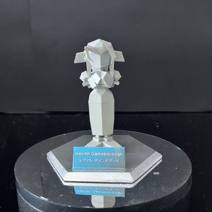 May include: White plastic figurine of a female character from a video game. The figurine is standing on a hexagonal base with a label that reads "Aerith Gainsborough" and "エアリス・ゲインズブール".