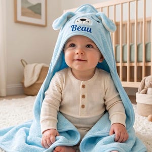 Peut inclure: Une serviette à capuche pour bébé bleu clair avec un motif de visage d'ours sur la capuche et le prénom Beau brodé en écriture cursive bleu foncé. La serviette est en tissu doux et pelucheux, portée par un bébé en body crème à manches longues.