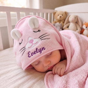 May include: A soft, light pink hooded baby towel featuring an embroidered cat face with closed eyes, whiskers, and a pink nose. The name Evelyn is embroidered in purple script below the cat face. The hood includes two small, plush cat ears.