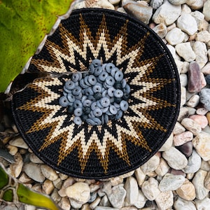 May include: A round, woven basket featuring a black, tan, and cream geometric starburst pattern. The centre of the basket is filled with a collection of small, circular, slate-grey beads with a matte finish and a central hole in each piece.