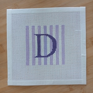K&ouml;nnte beinhalten: Ein quadratisches Stickleinen mit einem zentralen dunkelvioletten Buchstaben D vor einem Hintergrund aus abwechselnden hellvioletten und wei&szlig;en vertikalen Streifen. Das Leinen hat an allen vier Seiten wei&szlig;e Stoffbandr&auml;nder am Netzgewebe.