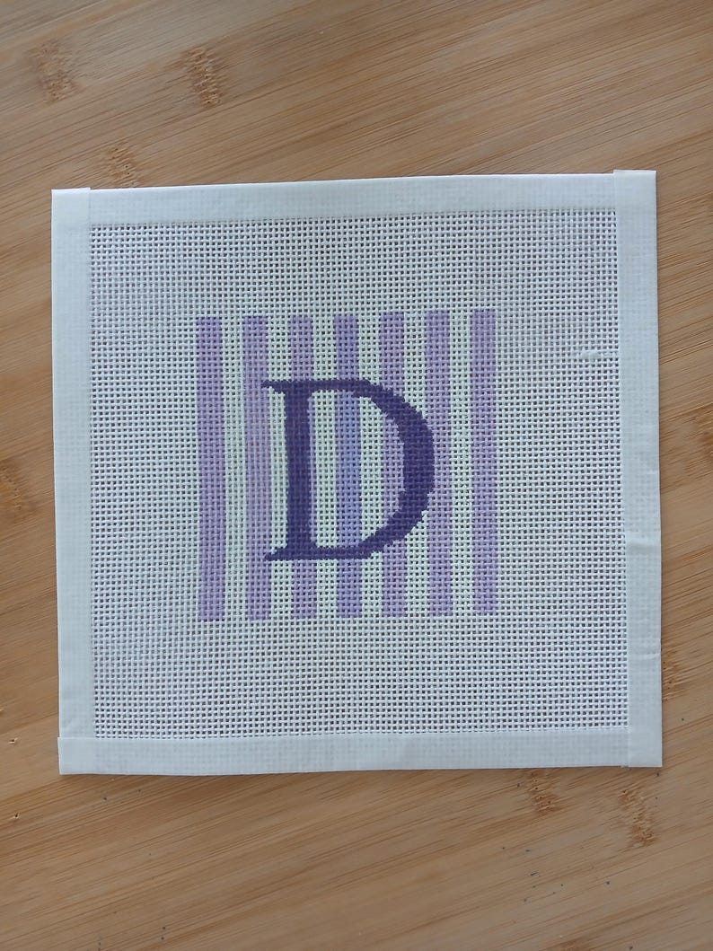 K&ouml;nnte beinhalten: Ein quadratisches Stickleinen mit einem zentralen dunkelvioletten Buchstaben D vor einem Hintergrund aus abwechselnden hellvioletten und wei&szlig;en vertikalen Streifen. Das Leinen hat an allen vier Seiten wei&szlig;e Stoffbandr&auml;nder am Netzgewebe.