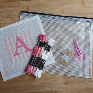 K&ouml;nnte beinhalten: Ein Stickset bestehend aus einer wei&szlig;en Leinwand mit einem rosa Buchstaben A und vertikalen Streifen, vier Stickgarnstr&auml;ngen in Rosa- und Wei&szlig;t&ouml;nen, einer kleinen rosa Schere, zwei Nadeln auf einer Karte und einer durchsichtigen Netztasche.