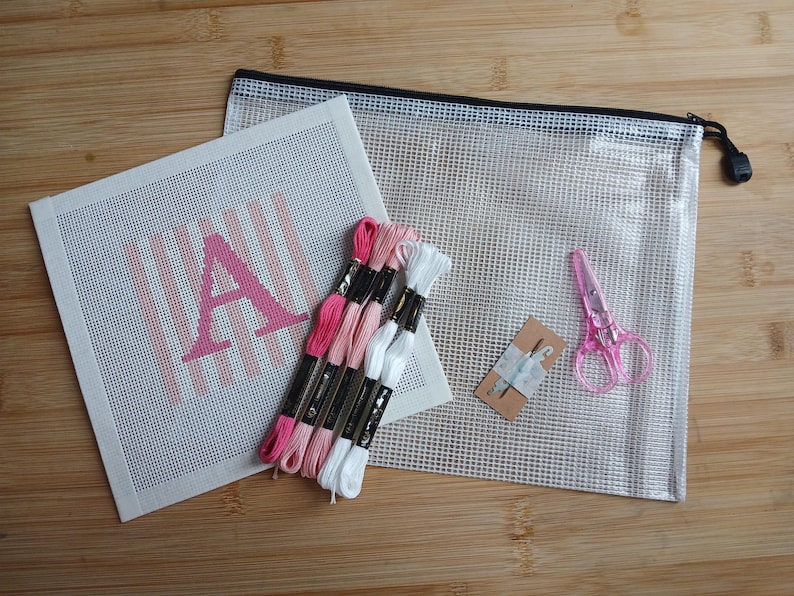 K&ouml;nnte beinhalten: Ein Stickset bestehend aus einer wei&szlig;en Leinwand mit einem rosa Buchstaben A und vertikalen Streifen, vier Stickgarnstr&auml;ngen in Rosa- und Wei&szlig;t&ouml;nen, einer kleinen rosa Schere, zwei Nadeln auf einer Karte und einer durchsichtigen Netztasche.