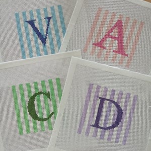 K&ouml;nnte beinhalten: Vier quadratische Stickleinw&auml;nde mit den gestickten Buchstaben V, A, C und D. Jeder Buchstabe ist &uuml;ber einem Hintergrund aus vertikalen Streifen in Hellblau, Rosa, Gr&uuml;n bzw. Violett zentriert, mit wei&szlig;en Stoffr&auml;ndern.