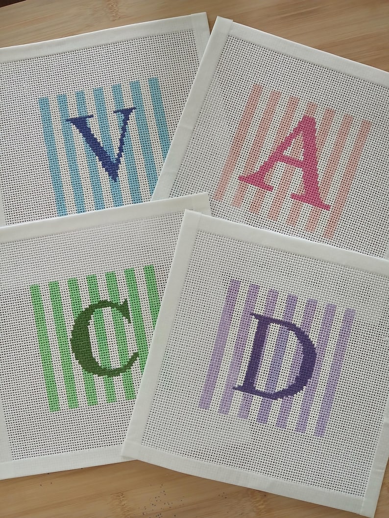 K&ouml;nnte beinhalten: Vier quadratische Stickleinw&auml;nde mit den gestickten Buchstaben V, A, C und D. Jeder Buchstabe ist &uuml;ber einem Hintergrund aus vertikalen Streifen in Hellblau, Rosa, Gr&uuml;n bzw. Violett zentriert, mit wei&szlig;en Stoffr&auml;ndern.