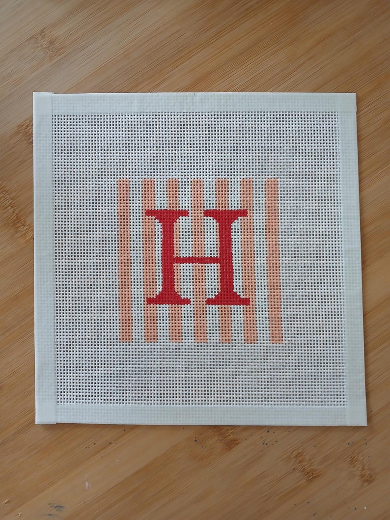 K&ouml;nnte beinhalten: Ein quadratisches Stickbild auf Stramin mit einem zentrierten, kr&auml;ftig roten Buchstaben H. Der Buchstabe liegt &uuml;ber einem Hintergrund aus vertikalen, hellpfirsichfarbenen Streifen. Der Stramin hat einen wei&szlig;en Klebebandrand an allen vier Seiten.