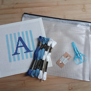 May include: A needlepoint kit featuring a square canvas with a blue striped pattern and the letter A, five skeins of blue and white embroidery floss, a small needle on a card, a pair of light blue scissors, and a clear mesh zippered storage pouch.
