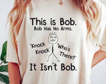 May include: A white t-shirt with a black graphic design that says "This is Bob. Bob Has No Arms." The graphic also includes a stick figure with a smile and the text "Knock Knock" and "Who's There?"  The text at the bottom of the graphic says "It Isn't Bob."