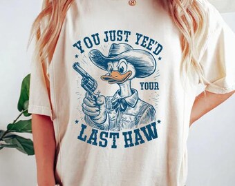 Op de afbeelding: Een crèmekleurig T-shirt met een cartoon eend die een cowboyhoed draagt en een pistool vasthoudt. De tekst op het shirt luidt "You just yeed your last haw".