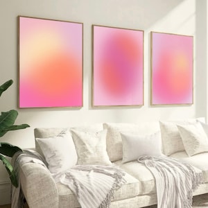 May include: Three framed abstract art prints with a gradient of pink, orange, and yellow colors. The prints are hanging on a white wall above a white couch with white throw pillows and a white blanket.