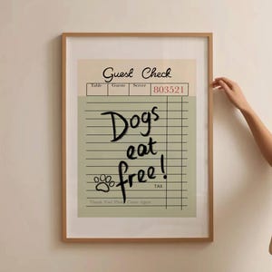 May include: A framed print featuring a vintage-style "Guest Check" design. The check reads "Dogs eat free!" in a playful font, with a paw print graphic. The frame is a light brown color.