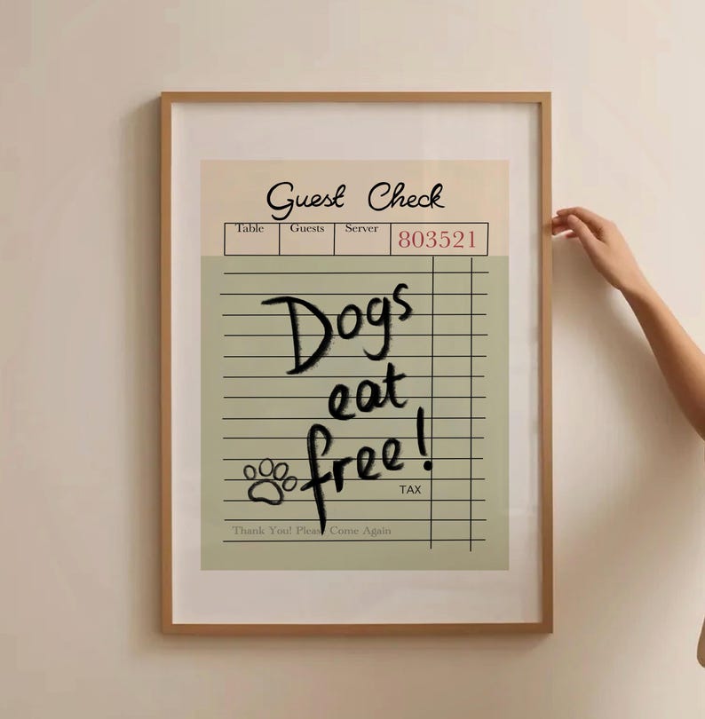 May include: A framed print featuring a vintage-style "Guest Check" design. The check reads "Dogs eat free!" in a playful font, with a paw print graphic. The frame is a light brown color.