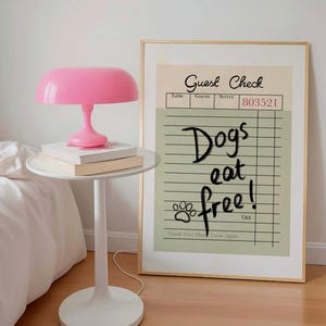 May include: A pink table lamp with a rounded shade sits on a white side table next to a framed print. The print reads "Guest Check" and "Dogs eat free!" in black text. Two books are stacked under the lamp.