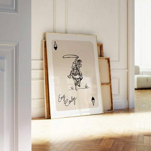 May include: A framed print of a playing card with a cowboy riding a horse and lassoing. The card is an Ace of Spades and the text "Easy Cowboy" is written below the image.