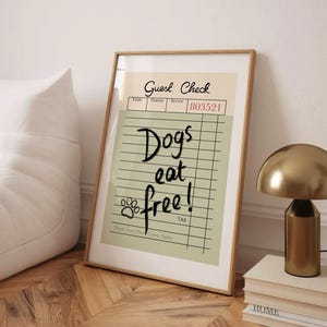 May include: Framed print with a tan and green background. The text "Guest Check" is at the top, with "Dogs eat free!" written in a handwritten style. A paw print is included. The frame is a light brown color.