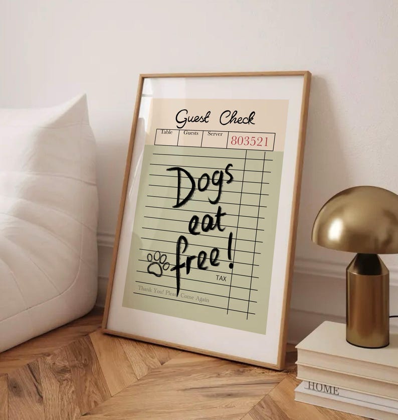 May include: Framed print with a tan and green background. The text "Guest Check" is at the top, with "Dogs eat free!" written in a handwritten style. A paw print is included. The frame is a light brown color.