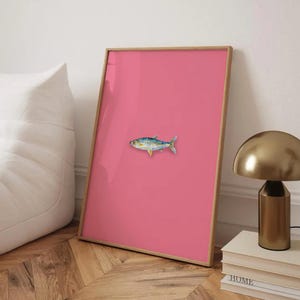 Puede incluir: Una impresión enmarcada de una pintura de acuarela de un pez sobre un fondo rosa. El pez es azul, verde y amarillo con el vientre blanco.