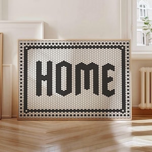 May include: A framed wall art piece displaying the word "HOME" in large, black block letters. The letters are composed of small, black hexagonal tiles, set against a background of white hexagonal tiles. The frame is a light brown colour.
