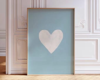 May include: A framed art print featuring a large white heart painted on a light blue background. The artwork is displayed in a gold-colored frame and leans against a white wall with decorative molding. The floor is made of wood.