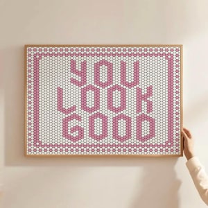 May include: A framed art print with the words "YOU LOOK GOOD" in pink, set against a white and pink hexagonal tile pattern. The artwork is in a light brown wooden frame.