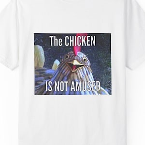 May include: White t-shirt with a graphic of a chicken and the text "The Chicken Is Not Amused." The image features a close-up of a chicken's face with a slightly open beak and a red comb. The shirt has a crew neck and short sleeves.
