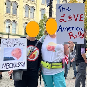 May include: Two people at a rally holding signs. One sign says "Grab em by the Neckussy" with a photo. The other sign reads "The Love America Rally". A building and other people are visible in the background.