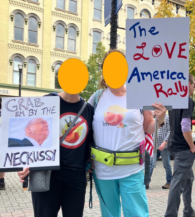 May include: Two people at a rally holding signs. One sign says "Grab em by the Neckussy" with a photo. The other sign reads "The Love America Rally". A building and other people are visible in the background.