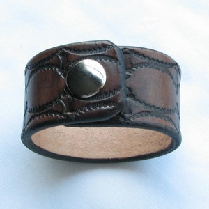 May include: A brown leather cuff bracelet with a silver snap closure. The bracelet has a decorative embossed pattern.