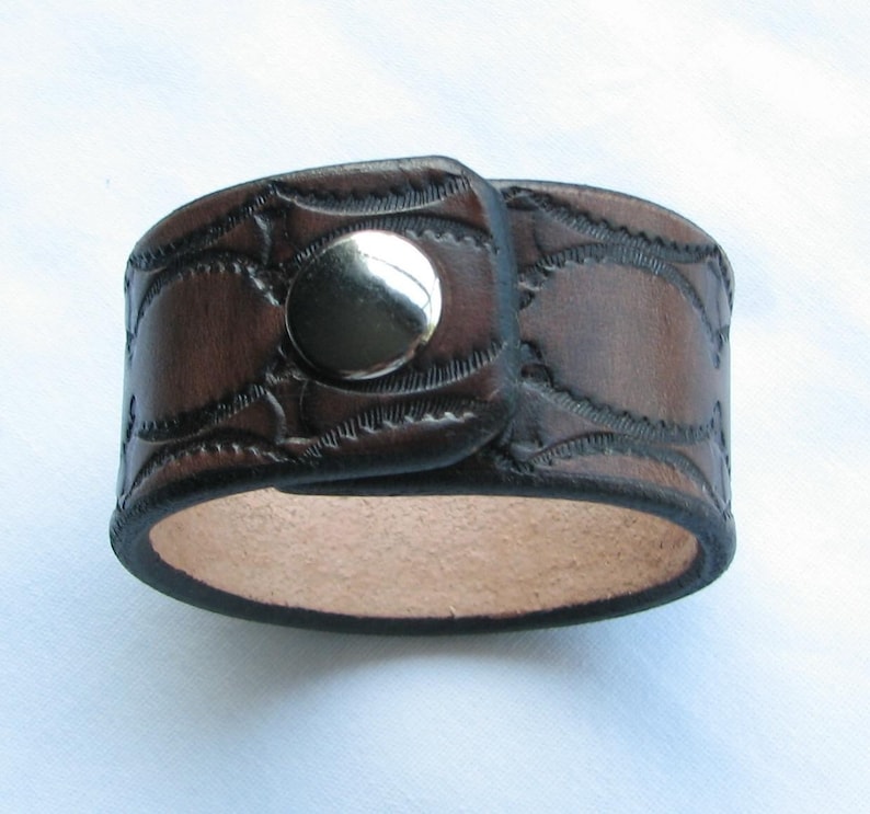 May include: A brown leather cuff bracelet with a silver snap closure. The bracelet has a decorative embossed pattern.