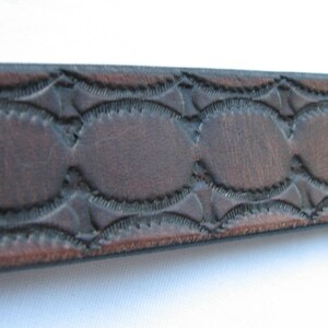 May include: Close-up of a brown leather belt with a decorative embossed pattern. The pattern features a series of oval shapes with a scalloped edge.