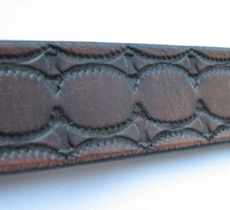 May include: Close-up of a brown leather belt with a decorative embossed pattern. The pattern features a series of oval shapes with a scalloped edge.