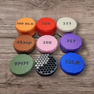 Puede incluir: Una colección de contenedores redondos y coloridos con etiquetas como "300 BLK", "556" y "223". Los contenedores son de varios colores, incluyendo naranja, rojo y morado. Un contenedor está lleno de pequeños objetos plateados.