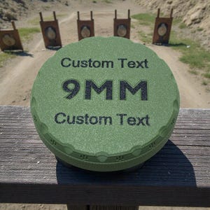 May include: A green, textured, round object with the text "Custom Text" above and below "9MM". The object is sitting on a wooden surface with a shooting range in the background.