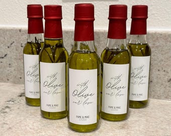 May include: Six small glass bottles with red lids filled with olive oil and sprigs of rosemary. Each bottle has a white label with black text that reads "with Olive our love" and "PAME & MIKE 5.1.2024".