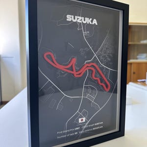 May include: A framed wall art piece featuring a dark gray map of the Suzuka race circuit with a raised red track outline. The text reads SUZUKA, First Grand Prix 1987, Circuit length 5.807 km, Number of laps 53, Race distance 307.471 km, and a Japan flag.