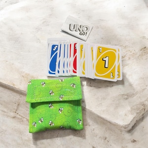 May include: A green fabric card holder with a bee pattern, next to a deck of Uno Go! cards. The cards are blue, red, and yellow with the number one on them. The Uno Go! card box is white with black text.