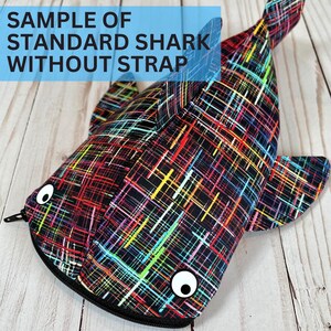 May include: A shark-shaped pouch with a zip closure, featuring a black base and a vibrant, abstract design of intersecting lines in red, yellow, green, and blue. The pouch has cartoon eyes and the text "SAMPLE OF STANDARD SHARK WITHOUT STRAP".