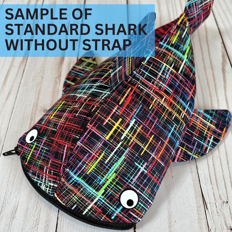 May include: A shark-shaped pouch with a zip closure, featuring a black base and a vibrant, abstract design of intersecting lines in red, yellow, green, and blue. The pouch has cartoon eyes and the text "SAMPLE OF STANDARD SHARK WITHOUT STRAP".