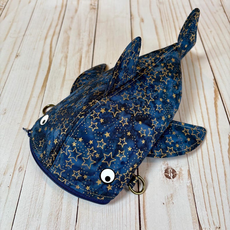 May include: A novelty fish-shaped pouch with a zip closure. The pouch is navy blue with a gold star pattern. It features two large, googly eyes and bronze-coloured metal rings. The fins and tail are also navy blue with the gold star pattern.