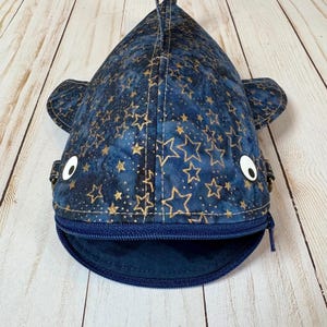 May include: A navy blue whale-shaped pouch with a zip closure. The fabric is decorated with gold star patterns. The pouch has white button eyes and small side fins. The zip is navy blue, and the pouch opens at the mouth.