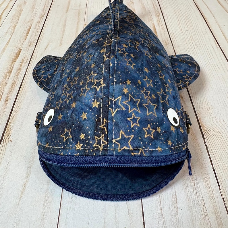 May include: A navy blue whale-shaped pouch with a zip closure. The fabric is decorated with gold star patterns. The pouch has white button eyes and small side fins. The zip is navy blue, and the pouch opens at the mouth.