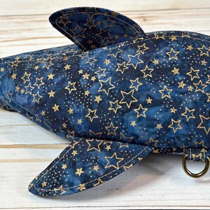 May include: A navy blue shark-shaped bag with gold star patterns. The bag has fins, a tail, and a white eye. It features a zip closure and a metal ring. The design is intended to look like a shark, made from fabric.