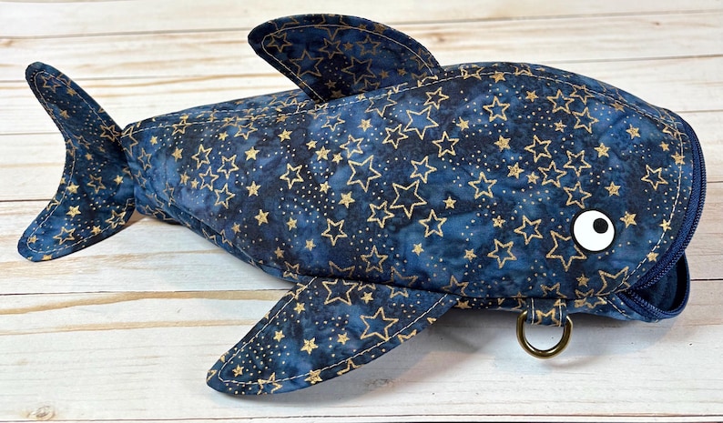 May include: A navy blue shark-shaped bag with gold star patterns. The bag has fins, a tail, and a white eye. It features a zip closure and a metal ring. The design is intended to look like a shark, made from fabric.