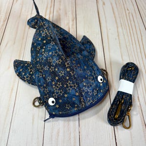 May include: A blue and gold star-patterned shark-shaped crossbody bag with a matching strap. The bag has a zip closure, white button eyes, and metal hardware. The strap is approximately 122 cm long.