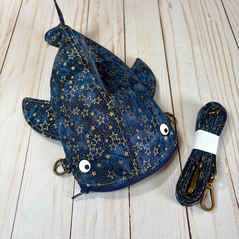 May include: A blue and gold star-patterned shark-shaped crossbody bag with a matching strap. The bag has a zip closure, white button eyes, and metal hardware. The strap is approximately 122 cm long.