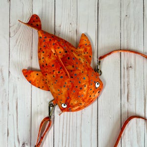 May include: Orange and blue spotted fish-shaped crossbody bag with a brown strap. The bag has two white eyes and a stitched mouth.