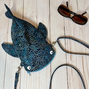 May include: A whimsical, blue and white polka-dotted whale-shaped crossbody bag with a zipper closure and a matching strap. The bag has white, cartoon-style eyes. Brown sunglasses rest on a light wood surface.