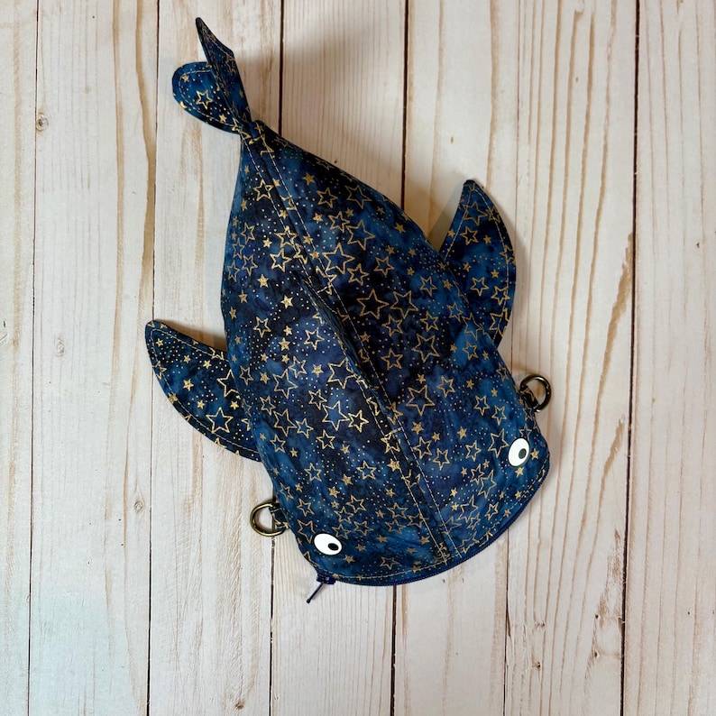 May include: A blue and gold star-patterned fish-shaped pouch with white button eyes and metal rings. The pouch is made of fabric and has a zip closure. The fish has fins and a tail.